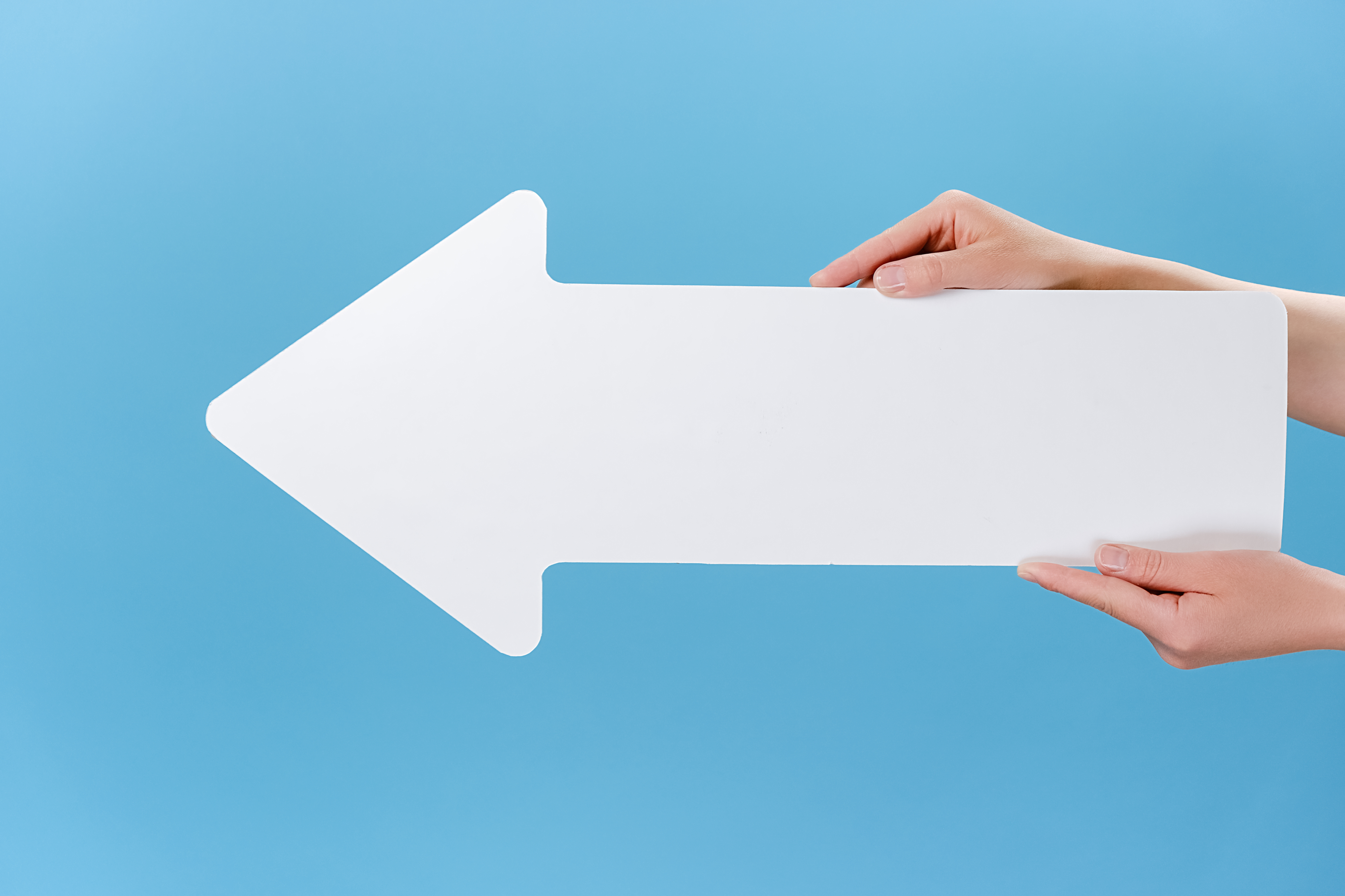 Close up cropped shot of hands holding a big white paper arrow, isolated on blue studio background wall