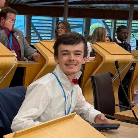 Bertie sits in holyrood at the youth parliament. He is wearing a white shirt, blue lanyard and smiling at he camera.