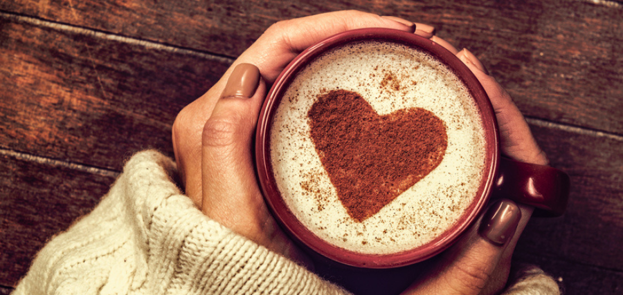 A cup of coffee, held by two hands around the cup, with a love heart latte art on top