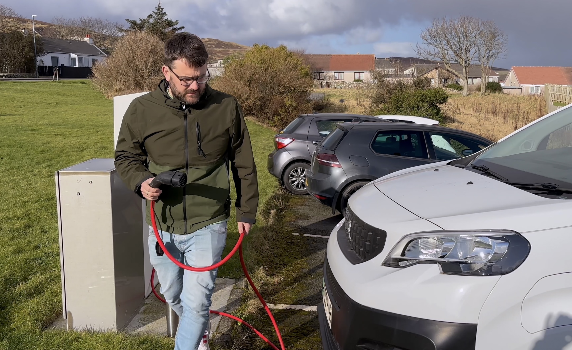 A person walks around the front of a white van holdinga plug, getting ready to charge the vehicle. 