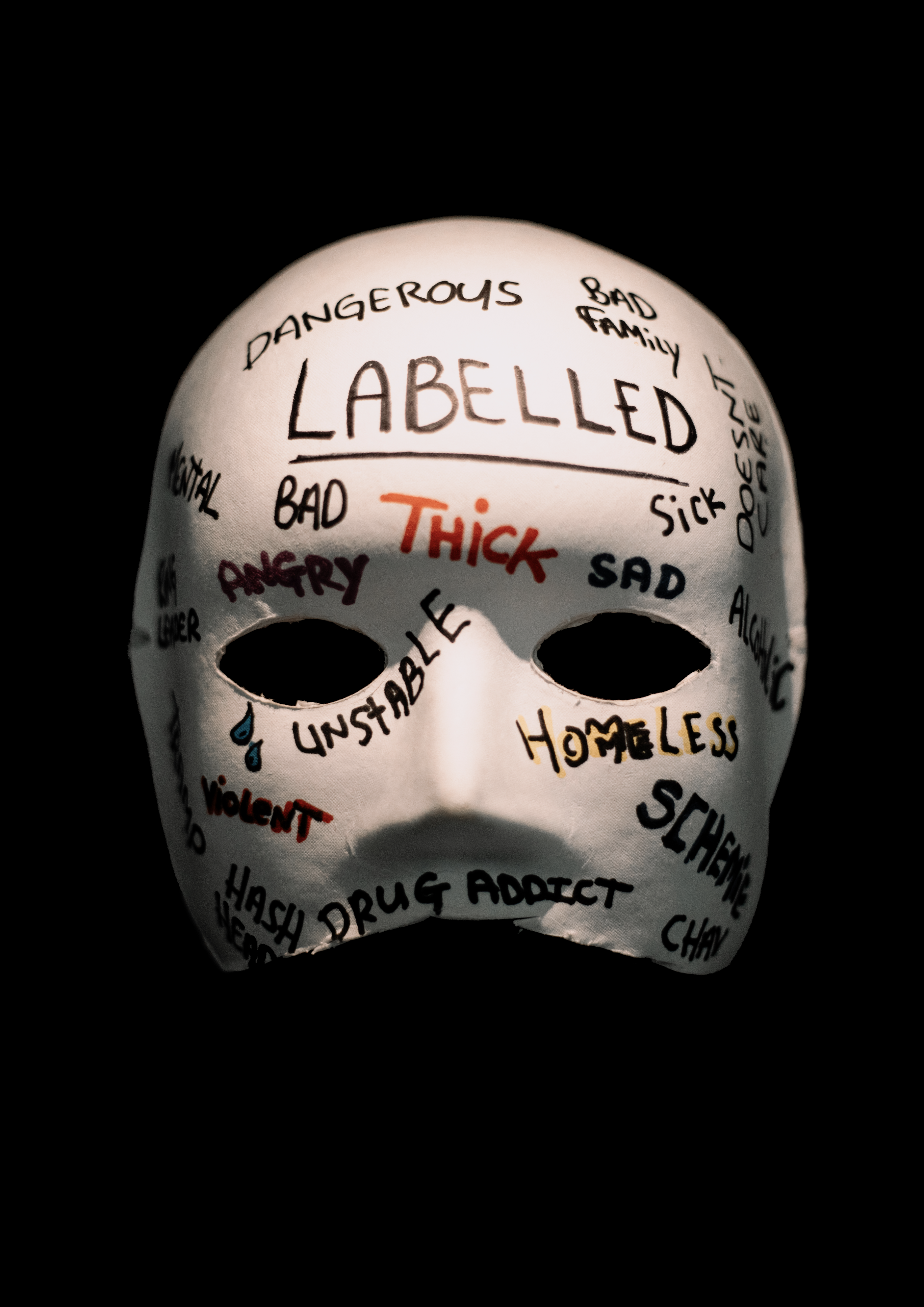 A white mask on a black background, across it is written words like 'labelled', 'thick', 'dangerous' 'bad' and 'homeless'. A small tear is drawn dripping from the masks eye.