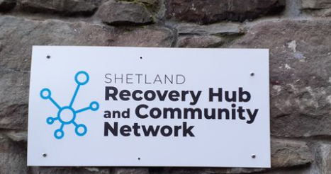 A white sign that states Shetland Recovery and Community Network. The logo is cyan circles that connect to a centre circle.