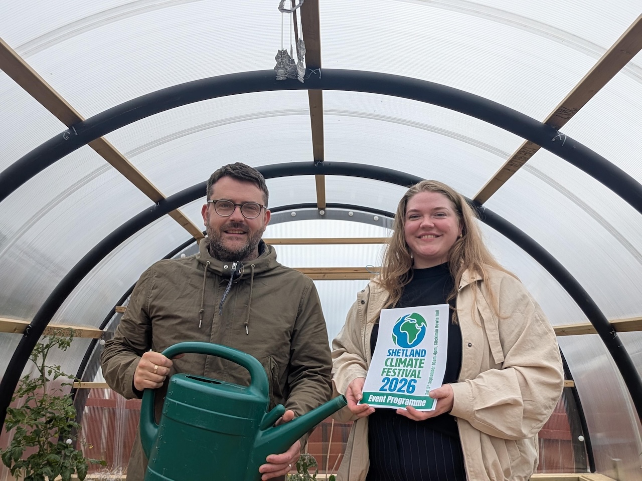 Anne & Elliot from the clmate change team hold the 2026 climate festival programme. They are standing in a polycrub