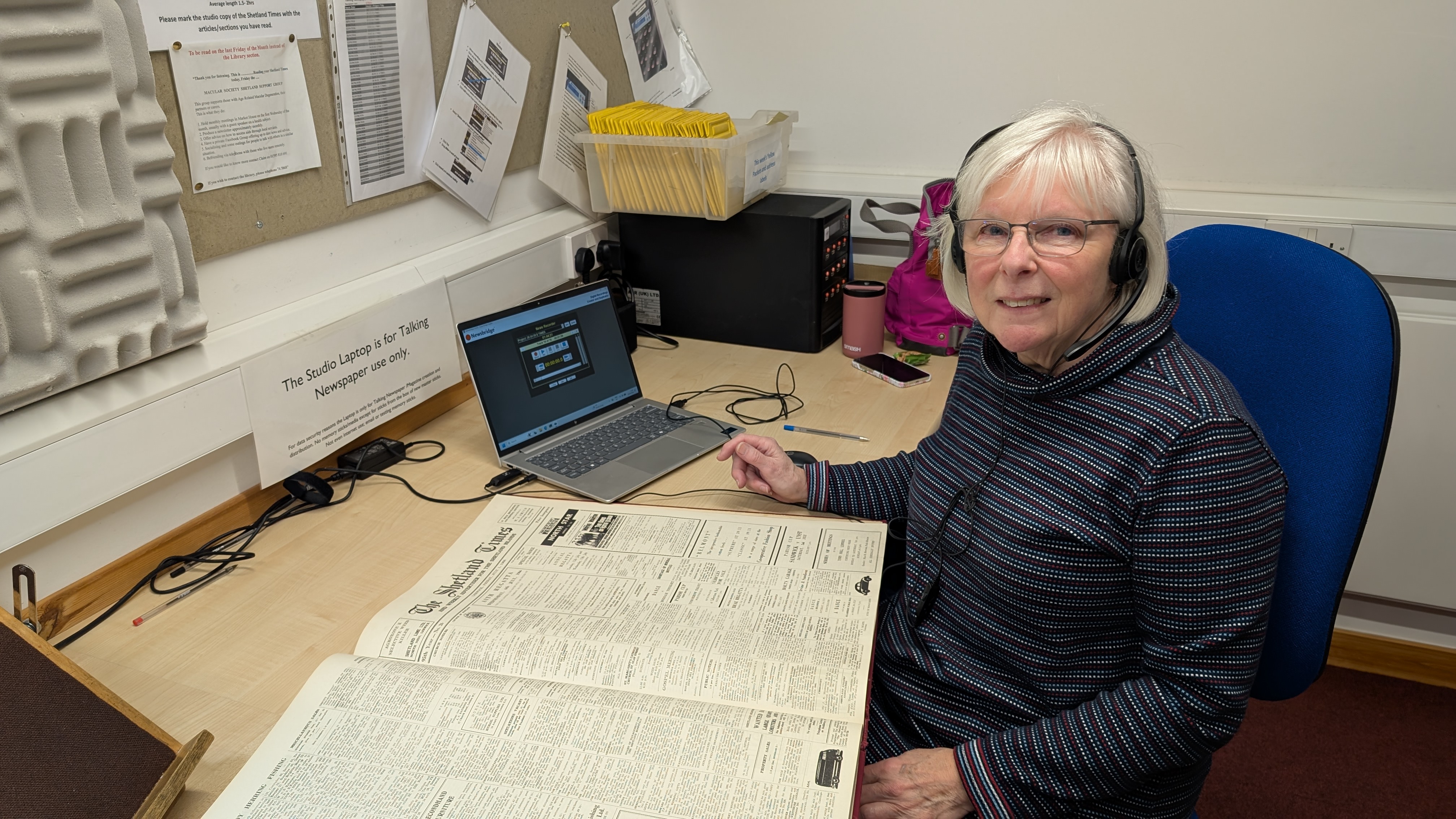 Jennie sits at a desk with an old copy of the Shetland Times spread in front of her. She is wearing headphones and looking at the camera